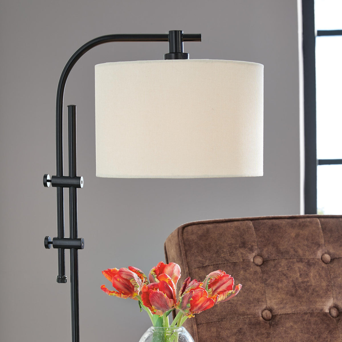 Table lamp with white shade and black base on a surface next to a vase with red tulips and a brown upholstered chair.
