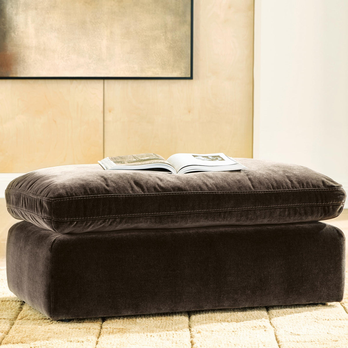 Brown ottoman with a magazine on top in a room with beige walls and a window.