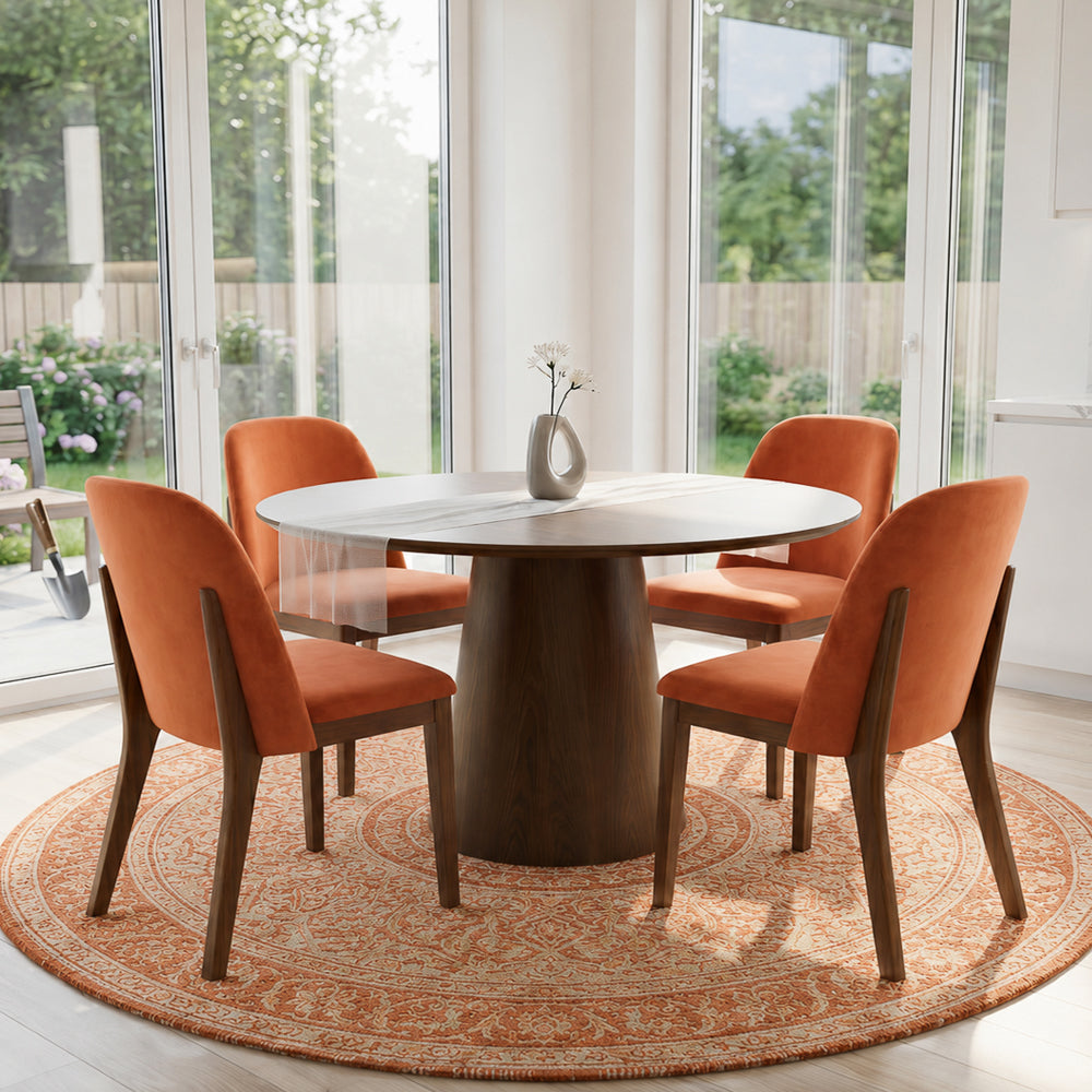 Four burnt orange dining chairs around a round marble-top table on a patterned rug by garden doors