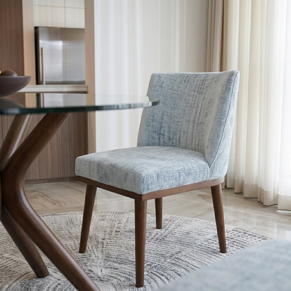 Grey upholstered dining chair beside a glass table, sheer curtains and kitchen behind