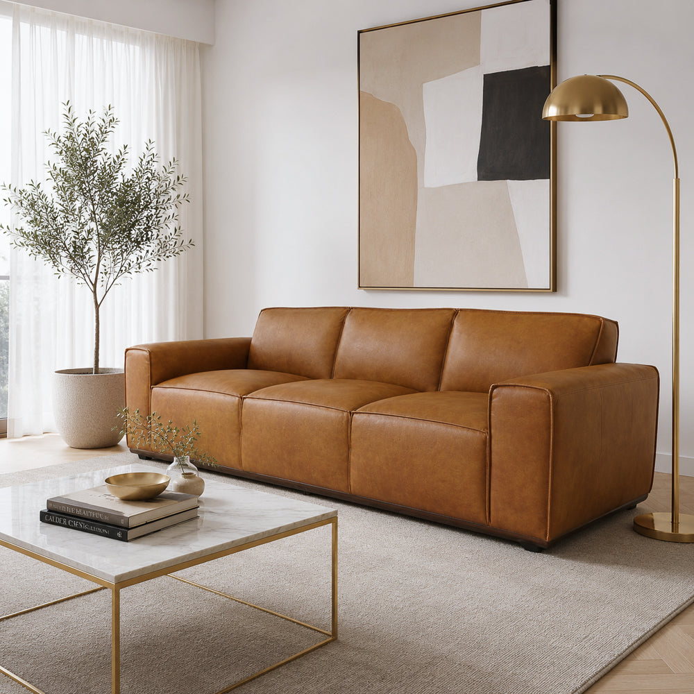 Angled living room view of the tan leather sofa beside a marble coffee table and sheer curtains