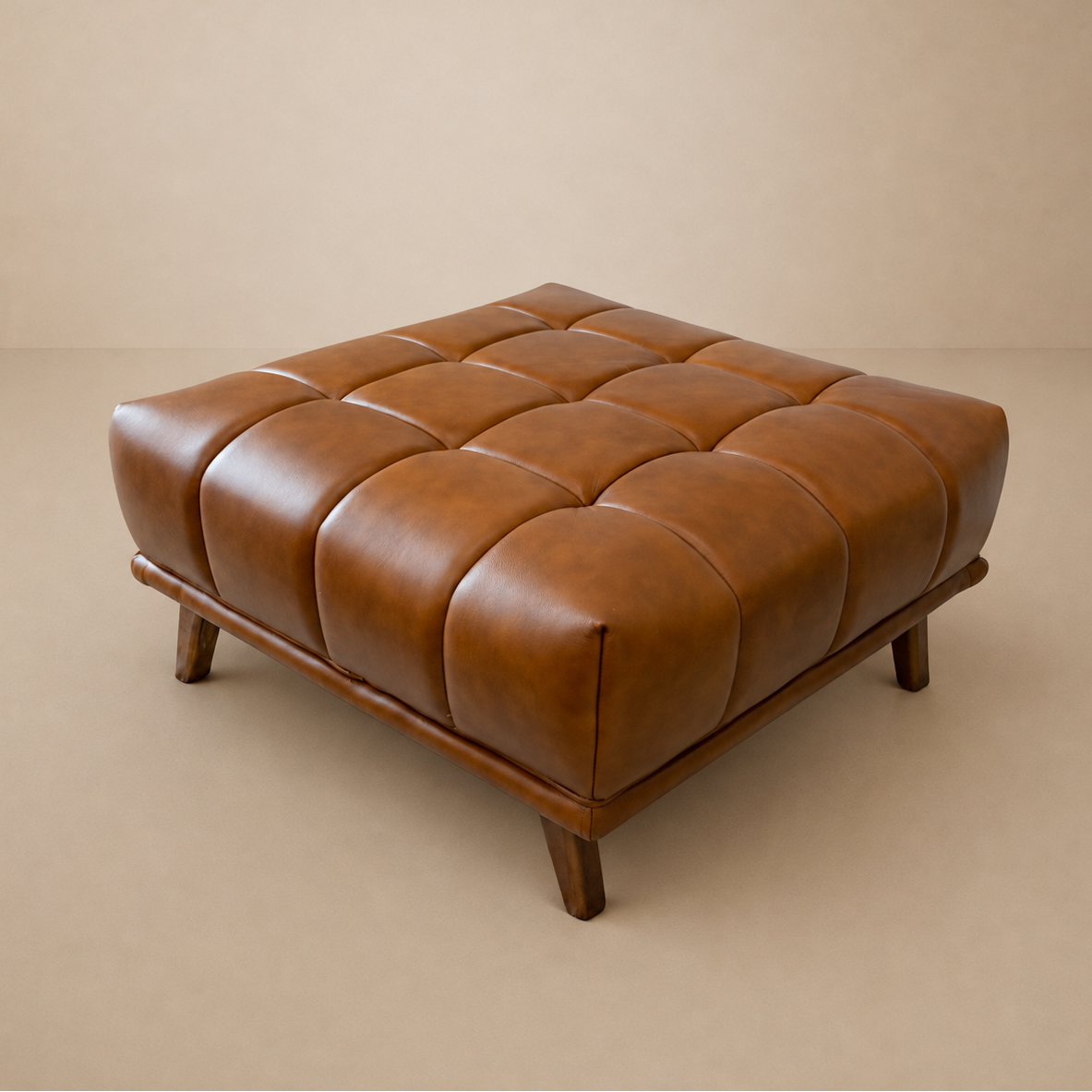 Cognac leather ottoman with tufted 16-square cushion grid and tapered wood legs, studio shot on a beige backdrop.