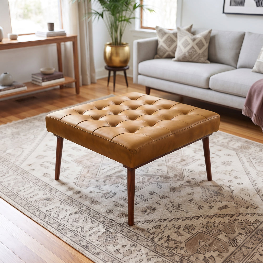 Three-quarter living-room shot of the tan leather ottoman on an ornate rug, gray sofa and gold planter in the background.