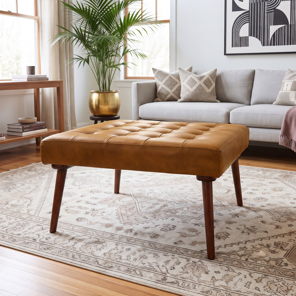 Front view of the tan tufted leather ottoman on a vintage rug, gray sofa and tall palm plant in the living room behind.