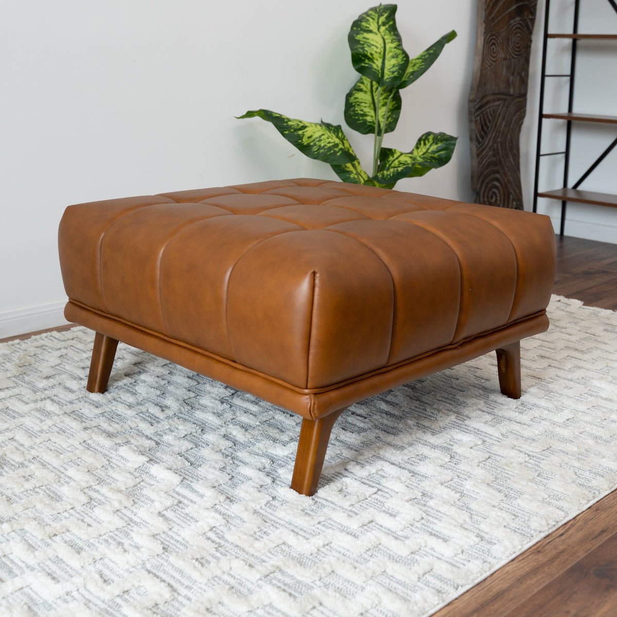 Side-front view of the leather ottoman on a textured rug, with a large variegated plant and wood sculpture behind.