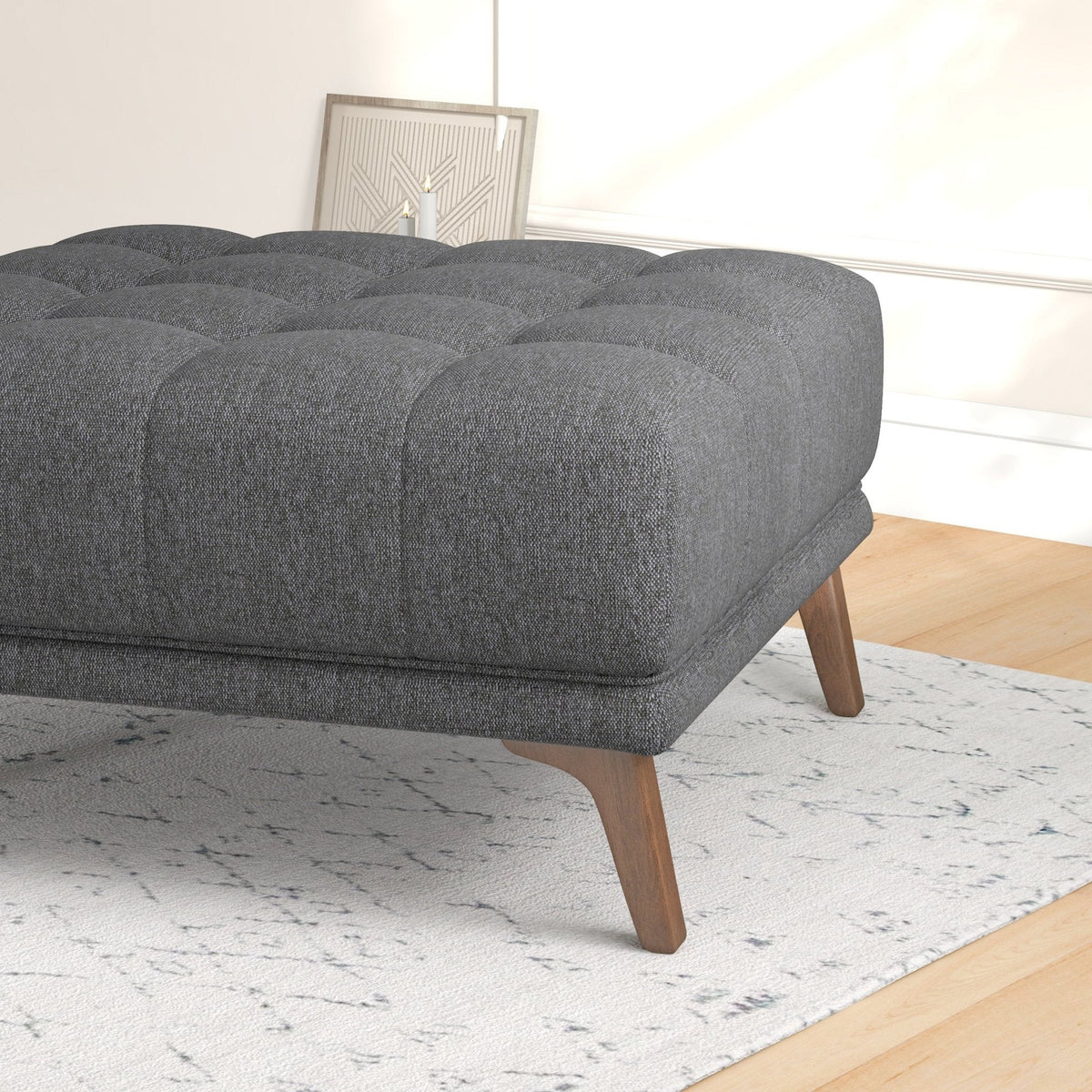 Close-up of the ottoman corner showing tufted dark gray fabric, thick cushion edge, and a tapered walnut-tone wood leg.