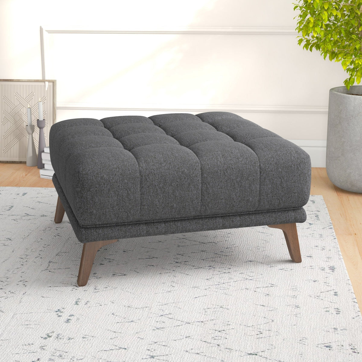 Side-angle view of the tufted dark gray ottoman on a textured rug, highlighting cushion depth and splayed wood legs.