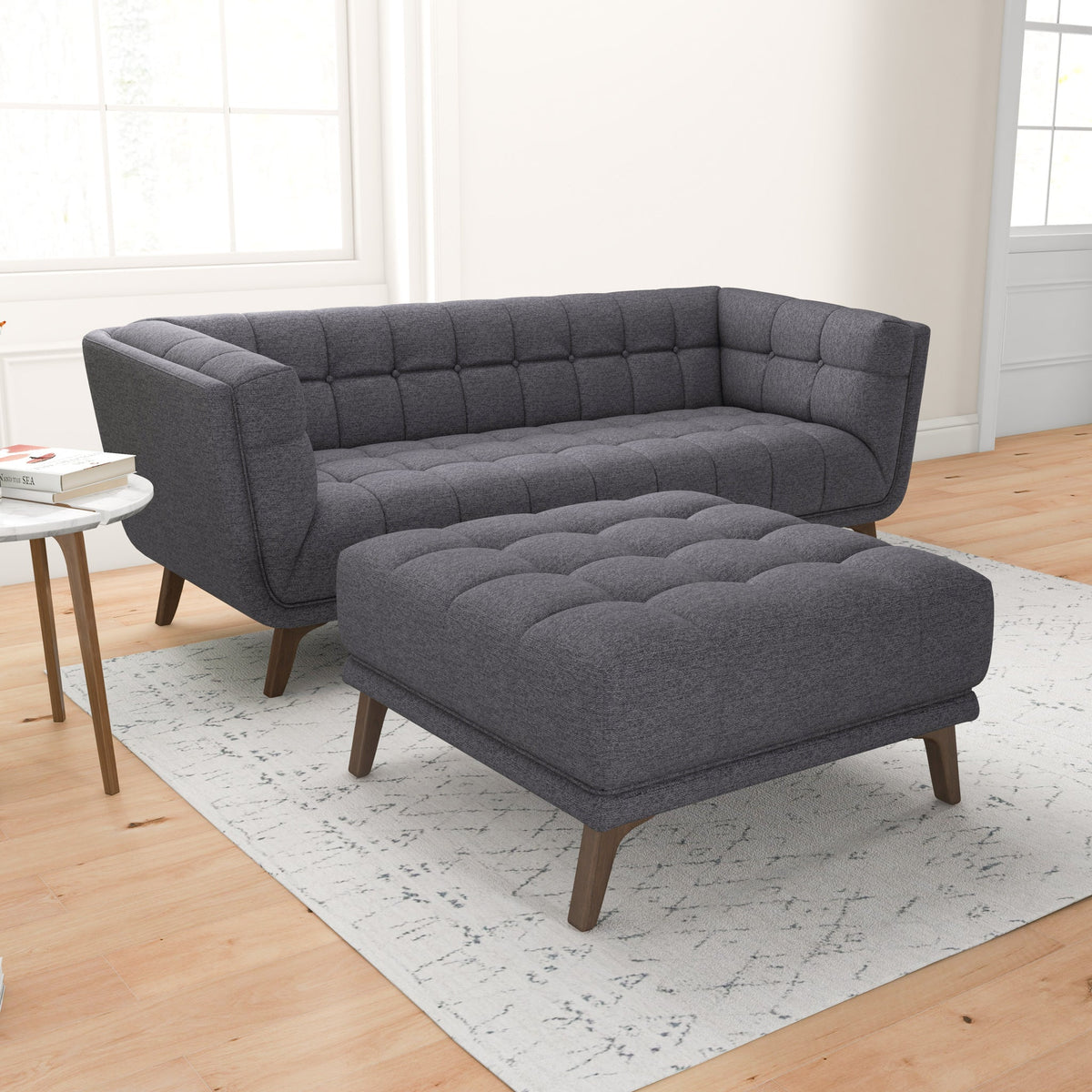 Corner view of a dark gray tufted sofa with the matching ottoman in front, two windows, and a small round side table.
