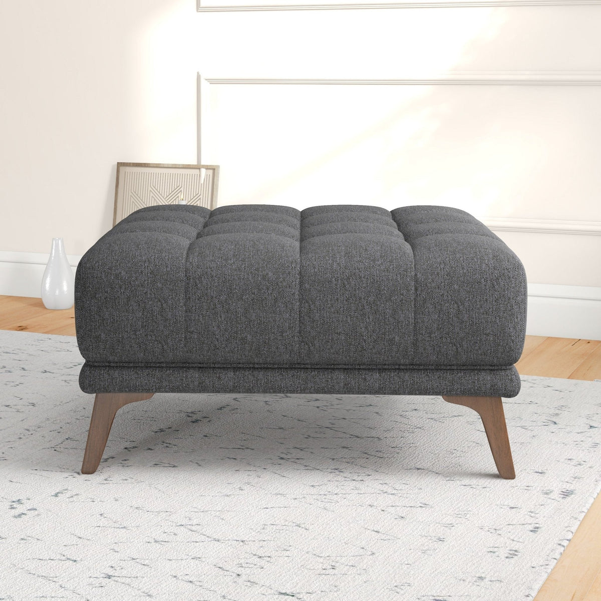 Front-facing view of the dark gray tufted ottoman on a light rug, with a white vase and framed art in the background.