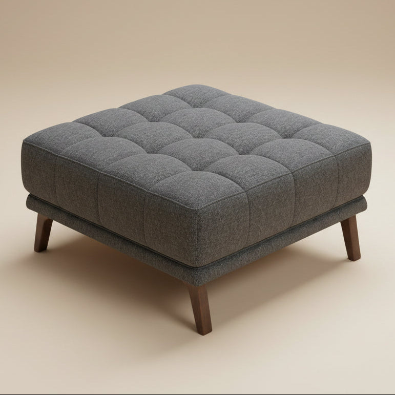Square dark gray tufted fabric ottoman with four tapered wood legs, isolated on a seamless beige studio background.