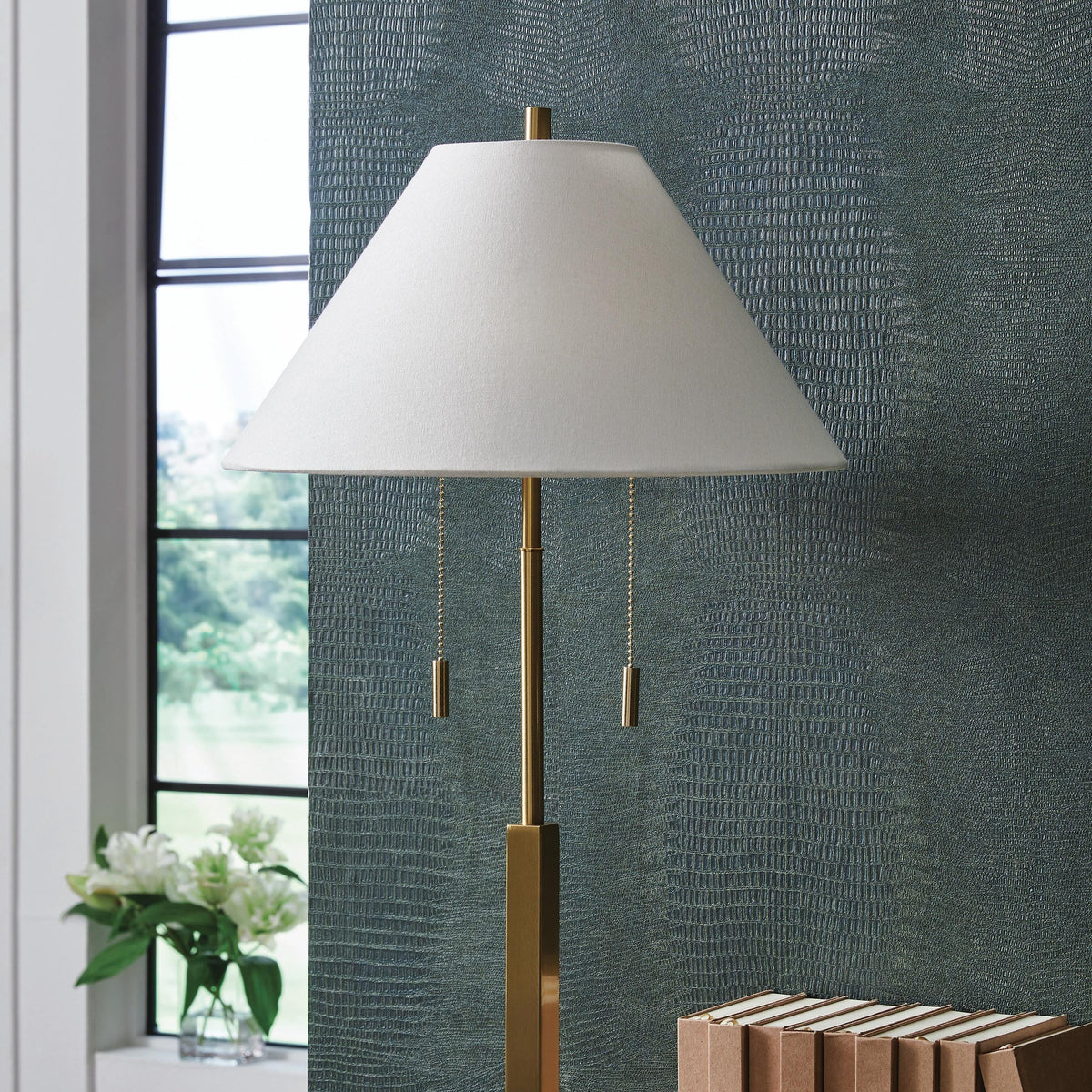 Gold floor lamp with white shade on a wooden floor next to books and apples.
