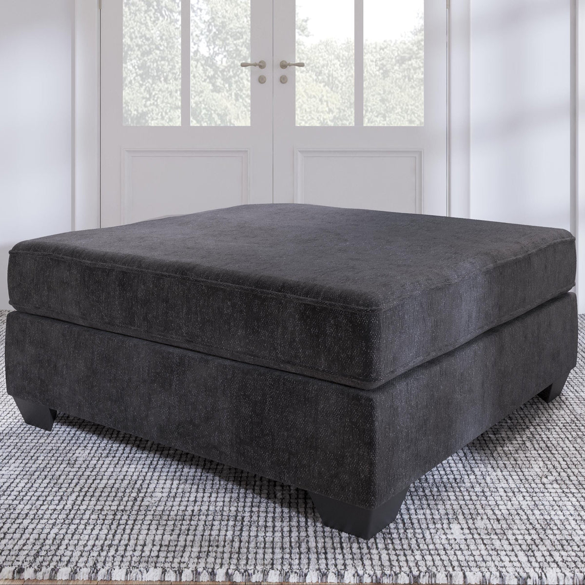 Gray ottoman in a room with white walls and a rug.