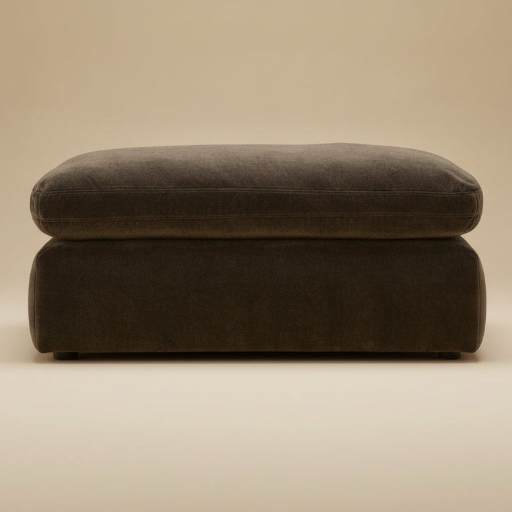 A dark brown upholstered ottoman with a thick cushioned top and rounded rectangular base, photographed against a warm beige studio background.