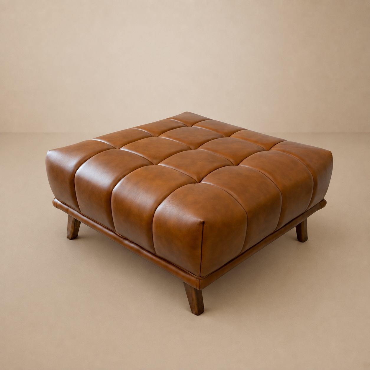 Cognac leather ottoman with tufted 16-square cushion grid and tapered wood legs, studio shot on a beige backdrop.
