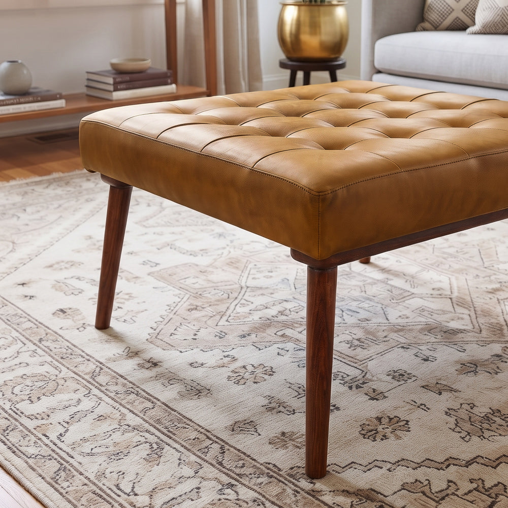 Low corner view of the tan leather ottoman highlighting tapered dark wood legs and tufted sides on a vintage rug.