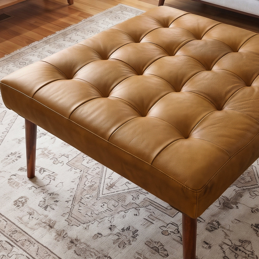 Close-up of the antique tan leather ottoman top, showing deep button tufting, grain, and the cushion's square edge.