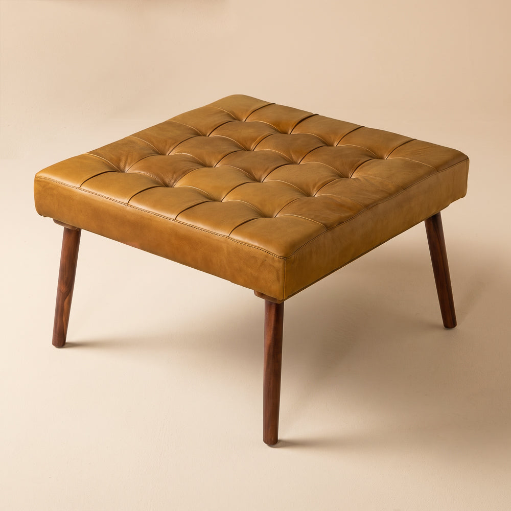Antique tan leather ottoman with tufted top and tapered wood legs, shown isolated on a plain beige backdrop.