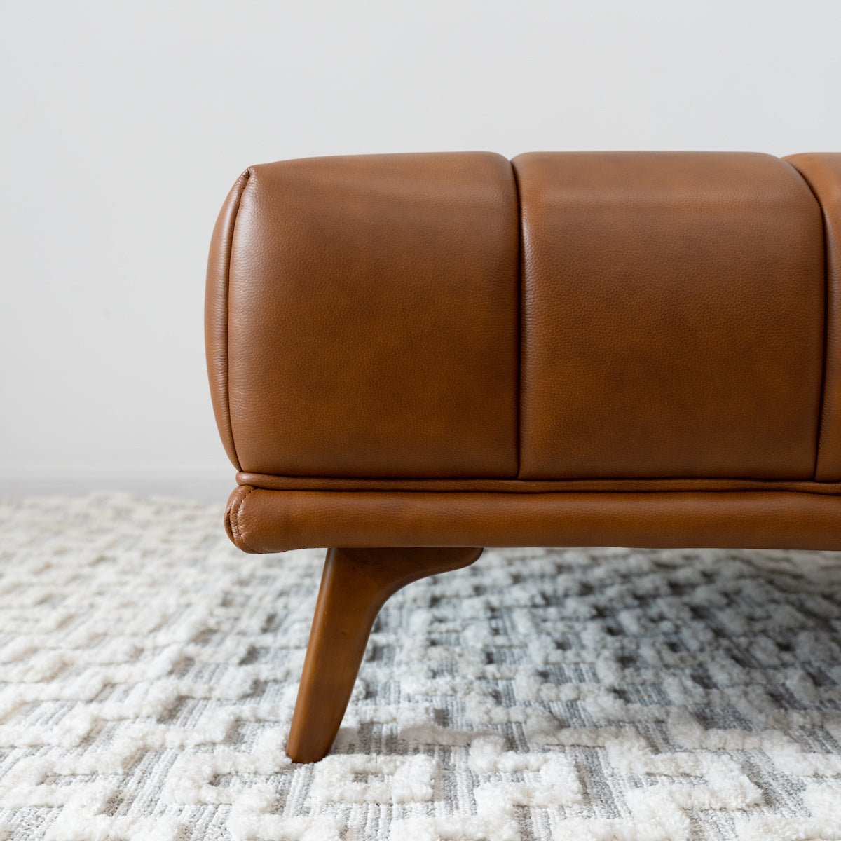 Close-up of the ottoman's rounded leather corner, vertical tufting seams, and a single tapered walnut-tone wood leg.