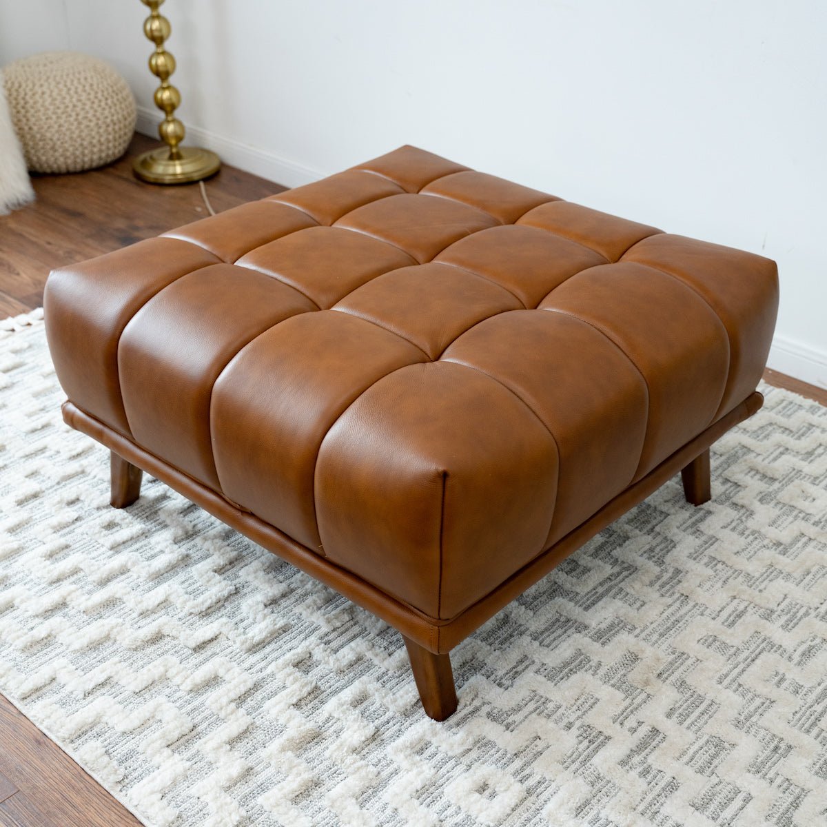 Angled view of the cognac tufted ottoman on a cream geometric rug, gold stacked-ball lamp and knit pouf behind.