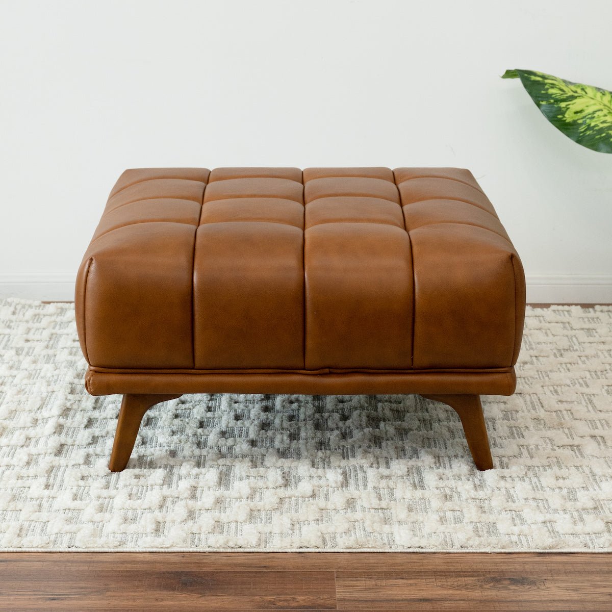 Front-facing view of the square cognac ottoman showing the 4-by-4 tufted top, centered on a cream patterned rug.