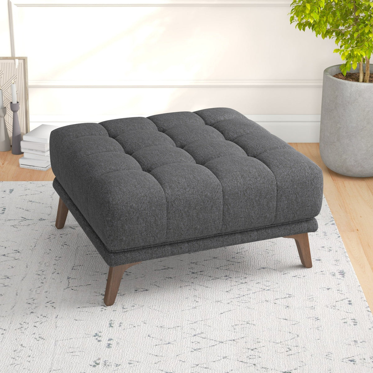 Dark gray tufted ottoman on a light patterned rug, angled toward a potted plant, books, and candles in a living room.